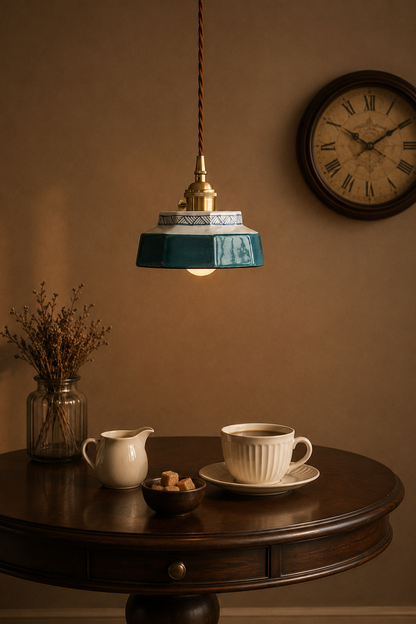 A vintage-style pendant light with a teal glass shade and brass fixture hangs over a dark wood table in a warm, rustic room. A white cup and saucer, a small cream pitcher, and a glass vase with dried stems sit on the table, with a round wall clock visible in the background.